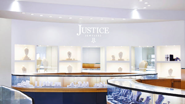 The Justice Jewelers store interior, with multiple glass-front cases and illuminated wall shelves. The wall prominently shows the brand name 'JUSTICE JEWELERS' above the display area.